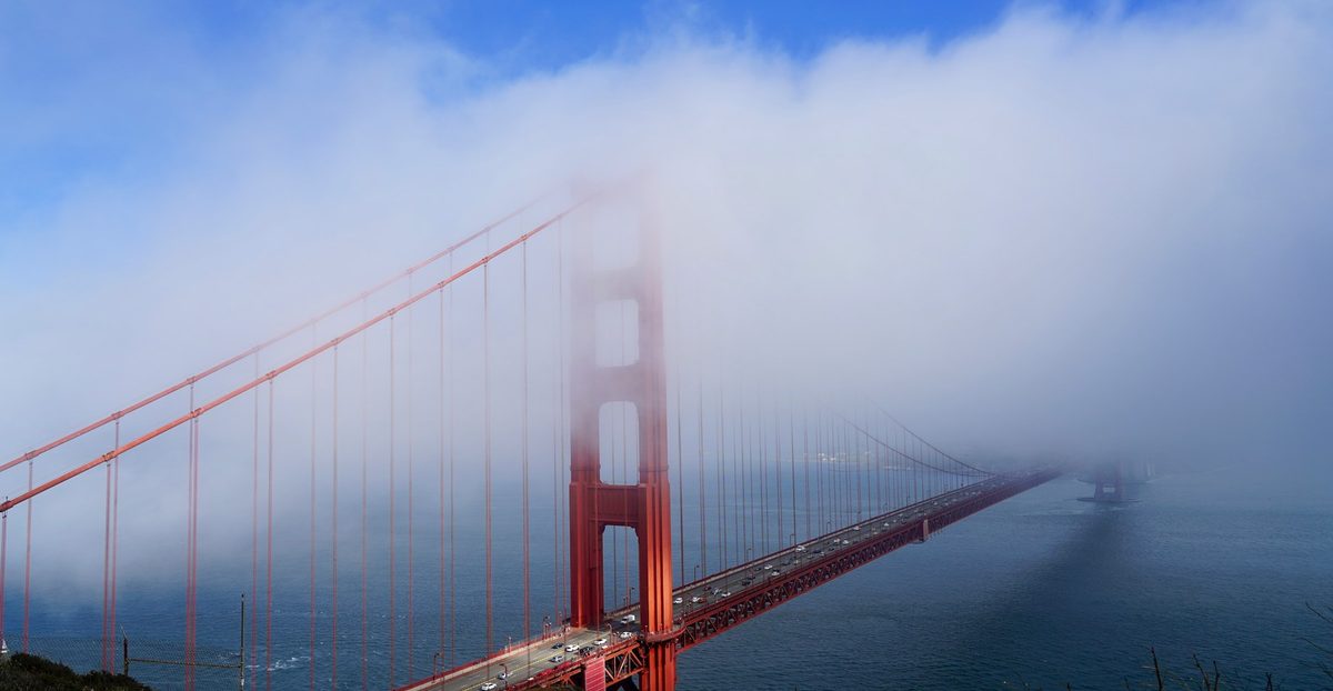 A red tower of the Golden Gate Bridge rising through a bank of fog over San Francisco Bay under a blue sky.