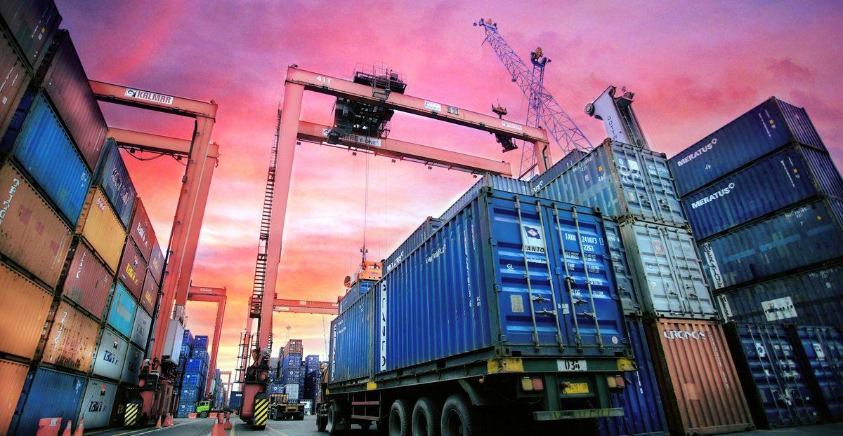 A truck hauling a blue shipping container between stacked containers and gantry cranes at a port under a pink sunset sky.