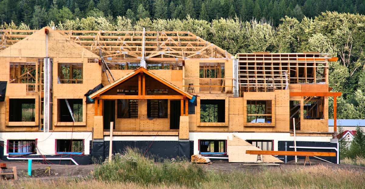 A multi-story wood-frame condominium building under construction, with exposed roof trusses and sheathed walls, in front of a forested hillside.
