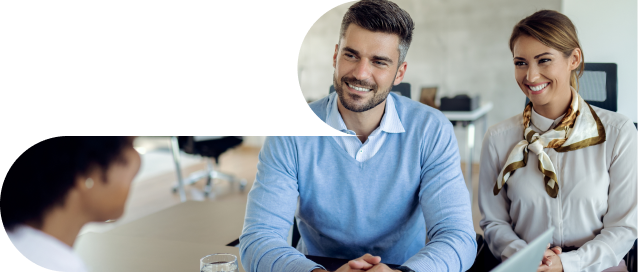 Two smiling professionals, a man in a light blue sweater and a woman in a white blouse and scarf, are seated across a table from another person (partially visible in the lower left) during what appears to be a business meeting or interview.