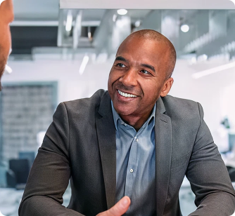 A smiling middle-aged man with short hair wearing a grey blazer and blue button-down shirt, looking toward another person during a conversation in a bright office.