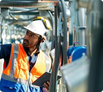 A male industrial worker wearing a white hard hat, safety glasses, and a high-visibility orange and blue vest. He is holding a digital tablet while inspecting complex machinery and piping in an industrial facility, looking upward with a focused expression.