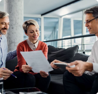 A diverse group of three professionals in a modern office lounge. A woman in an orange sweater and a man with a grey beard sit on a sofa, smiling as they review a document together. Opposite them, a man in a white shirt and glasses gestures while holding a digital tablet, engaging them in a collaborative discussion.