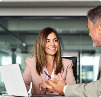 A smiling woman with long brown hair wearing a pink blazer sits at a desk with a laptop. She is looking toward a male colleague (partially visible in the foreground) who is gesturing with a pen during a professional consultation or interview in a bright, glass-walled office.