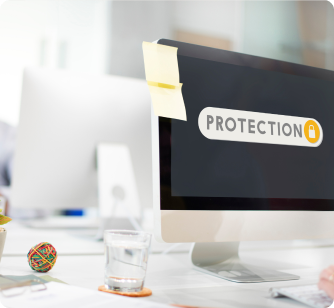 A computer monitor on a white office desk displaying a "PROTECTION" graphic with a yellow lock icon. A yellow sticky note is attached to the top of the monitor.