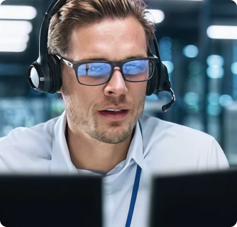 A focused customer support agent wearing a headset and glasses works at a computer in a modern office with softly lit screens in the background.