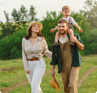 A happy family of three walks through a sun-drenched grassy field. The father wears overalls and carries a young toddler on his shoulders, while the mother walks beside them wearing a sun hat. They are surrounded by green trees under a soft, golden light.