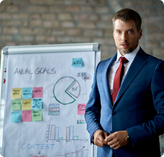 A businessman in a blue suit standing next to a whiteboard covered in charts, sticky notes, and project goals.