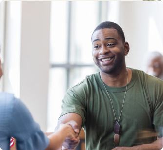 A smiling man wearing a green t-shirt and dog tags shakes hands with another person. The setting is a brightly lit indoor space with large windows in the background.