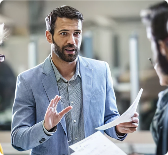 A man in a light blue blazer gestures with his hand while holding a stack of papers. He appears to be leading a conversation or presentation in a busy office environment.