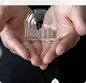 Two cupped hands gently hold a transparent glass dome containing stacked coins arranged like a city skyline, symbolizing financial protection or savings.