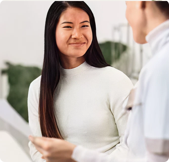 woman with long dark hair smiles warmly while looking at someone off-camera. She is wearing a white turtleneck sweater in a minimalist, modern room.
