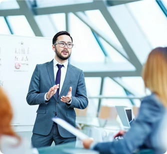 A professional man in a grey suit and glasses giving a presentation in a modern, bright office space with large geometric windows.