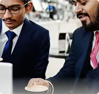 Two businessmen in suits having a casual professional conversation while holding cups of coffee in a bright office environment.