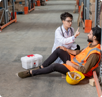 A female doctor in a white lab coat providing first aid to an injured warehouse worker sitting on the floor in a safety vest.