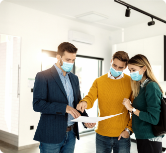 A real estate agent and a couple, all wearing face masks, examine a floor plan inside a bright, modern apartment.
