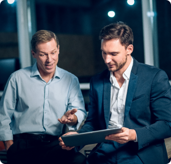 Two men in professional clothing sit together and review information on a digital tablet during a meeting.
