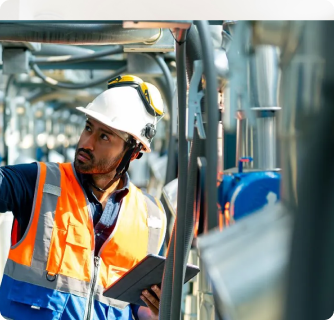 A male industrial worker wearing a white hard hat, safety glasses, and a high-visibility orange and blue vest. He is holding a digital tablet while inspecting complex machinery and piping in an industrial facility, looking upward with a focused expression.
