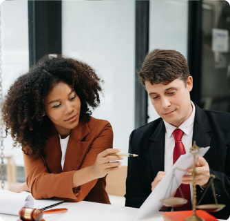 A female lawyer in a brown blazer points to a document held by a male colleague in a suit.