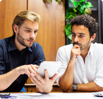 Two men sitting at a wooden desk, focused intently on a digital tablet held by the man on the left. They appear to be collaborating on a project in a professional office setting.