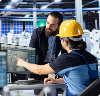 A man in a suit and a worker in a yellow hard hat collaborate while looking at a technical 3D model on a computer screen in a factory.