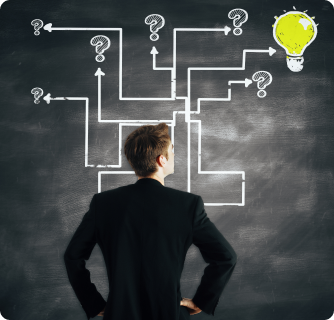 A businessman standing with his back to the camera, looking at a chalkboard filled with question marks and winding paths. One path leads to a glowing yellow lightbulb, symbolizing a successful idea.
