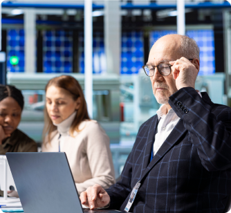 A senior male professional wearing glasses working on a laptop with colleagues in the background.