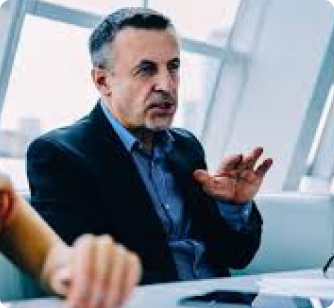 A middle-aged man in a dark blazer and blue shirt gestures with his hands while speaking during a professional meeting.