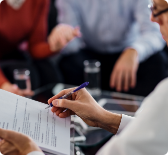 A close-up of a person’s hand using a pen to point at specific clauses in a document during a meeting. Other participants are blurred in the background, emphasizing the detailed scrutiny of a contract.