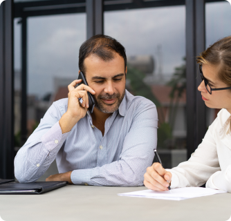 A man in a light grey shirt talks on his mobile phone while a woman in a white blouse takes notes beside him.