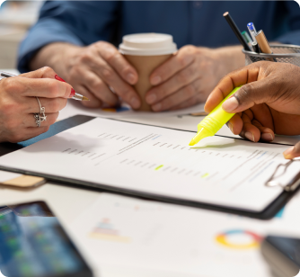 A close-up of hands at a meeting; one person uses a bright yellow highlighter on a document while another holds a pen.