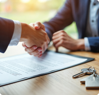 Two business people are sealing a deal, shaking hands over a table with a contract and house keys. It signifies the successful completion of a real estate or major business transaction.
