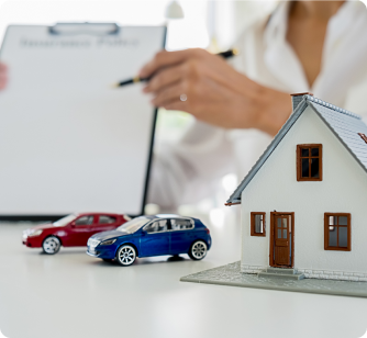 A miniature model of a house and two toy cars sit in the foreground, with the blurred background showing an insurance agent holding a clipboard and pen.