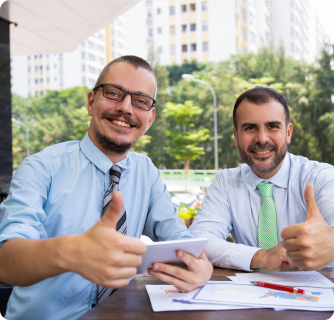 Two businessmen sitting outdoors giving a thumbs-up while holding a digital tablet.