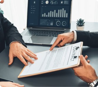 Two people in business attire sitting at a desk; one points to a "CONTRACT" document on a clipboard while a laptop in the background shows data dashboards.