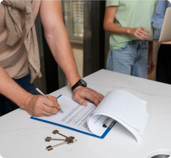 A person leans over a white counter to sign a contract on a clipboard next to a set of house keys. In the background, other people stand with a laptop, signifying a real estate closing or rental agreement.