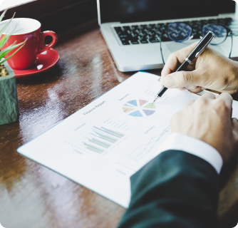 An over-the-shoulder view of a person in a suit reviewing a printed finance report with a pie chart, with a red coffee mug and a laptop on a wooden desk.