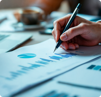 A close-up view of a hand holding a black pen and reviewing a financial report with blue bar charts and pie graphs.