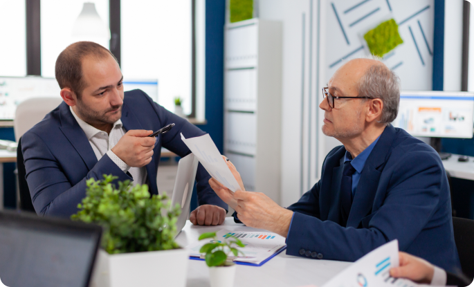 Two businessmen in suits sitting at a desk discussing a document in a modern office setting.