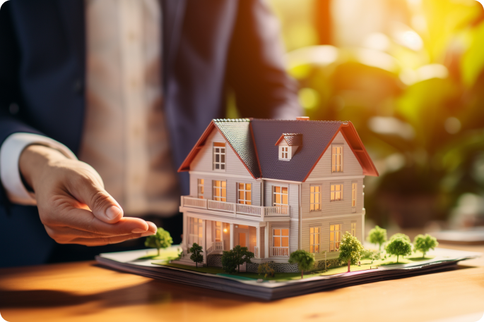 A man in a suit gestures toward a detailed architectural model of a two-story white house sitting on a stack of blueprints.