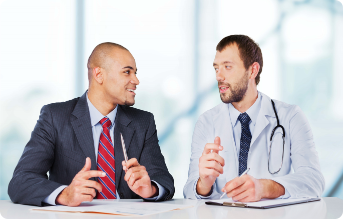 A professional in a striped tie and grey suit speaks with a doctor in a white coat who is holding a pen and a clipboard.