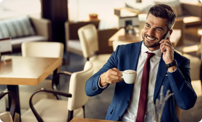 A smiling man in a blue suit and red tie sitting in a brightly lit cafe. He is holding a cup of coffee in one hand and talking on a mobile phone with the other.