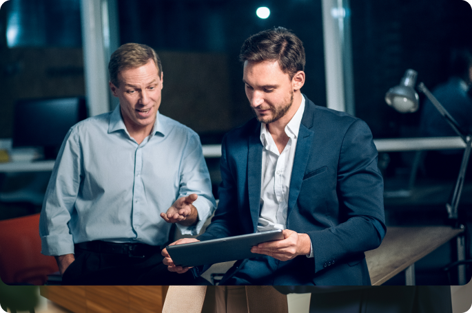 Two businessmen in an office at night; one sits and gestures toward a tablet screen while the other looks on with an engaged expression.