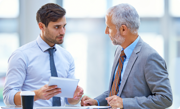 A younger businessman and an older executive discussing information on a tablet in a bright, modern office.