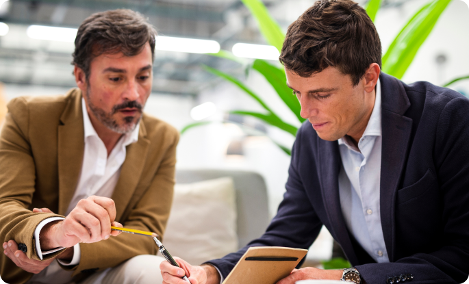 Two men in professional attire sit together closely reviewing and pointing at notes in a notebook with a pencil.