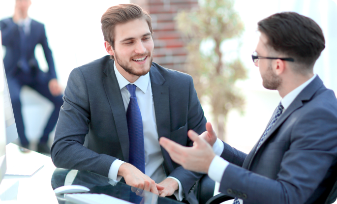 A smiling businessman in a grey suit and blue tie engaging in a friendly conversation with a colleague. They are sitting at a glass desk in a bright, modern office environment.