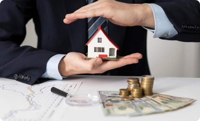 A professional in a dark suit holds a small white model house with a red roof in the palm of one hand while sheltering it with the other hand. The scene includes a desk with a line graph, a magnifying glass, stacks of coins, and US currency, illustrating real estate investment or home insurance.