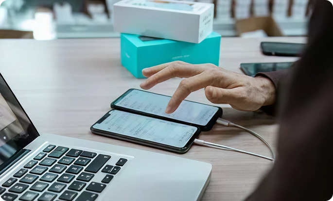 A person’s hand scrolls through data on two smartphones side-by-side on a desk, next to an open laptop and product boxes.