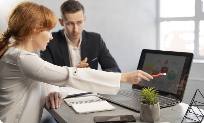 A woman with red hair points at a complex flowchart on a laptop screen while explaining a process to a male colleague in a bright office.
