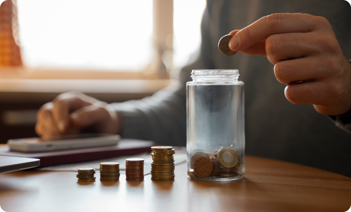 A person placing a coin into a glass jar that is partially filled with various coins. Stacks of coins of different heights sit on the table nearby, symbolizing savings and financial planning.